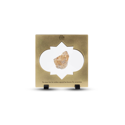 A natural stone is suspended in the middle of a square bronze frame with a traditional middle eastern inlay that is set on two little black square stands. On the top of the frame is the "MyEvven" logo that says Temple Mount Stones below it. On the bottom of the frame, it reads "The stone that the builders rejected has become the cornerstone".
