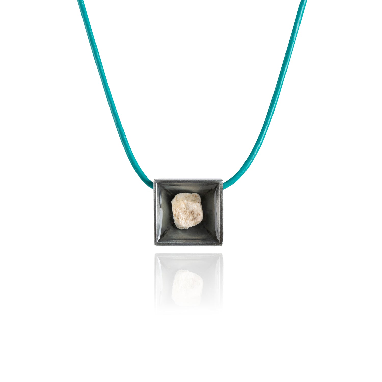 A small natural tan stone sitting in the middle of a square shaped metal pendant in a dark silver color. The pendant is hanging on a turquoise leather necklace.