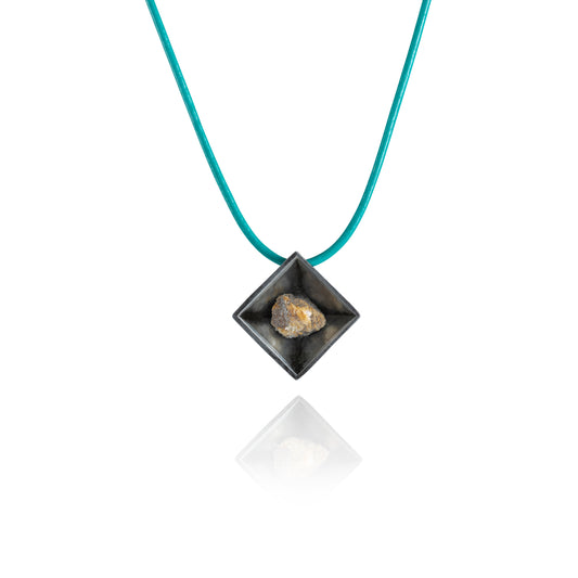 A small natural multicolored stone sits in the middle of a diamond shaped metal pendant in a dark silver color. The pendant is hanging on a turquoise leather necklace.