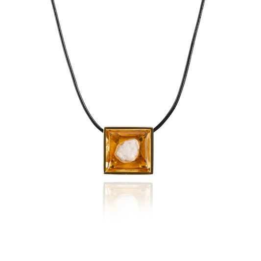 A small natural tan stone sitting in the middle of a square shaped metal pendant in a gold color. The pendant is hanging on a black leather necklace.